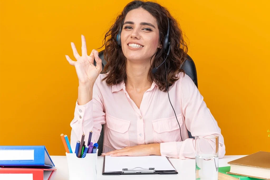 Asistente virtual para startups con auriculares haciendo el gesto de OK, sonriendo en un escritorio de oficina.