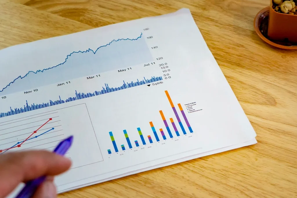 Hand holding a pen analyzing a printed financial growth report with stock charts and bar graphs on a wooden table.