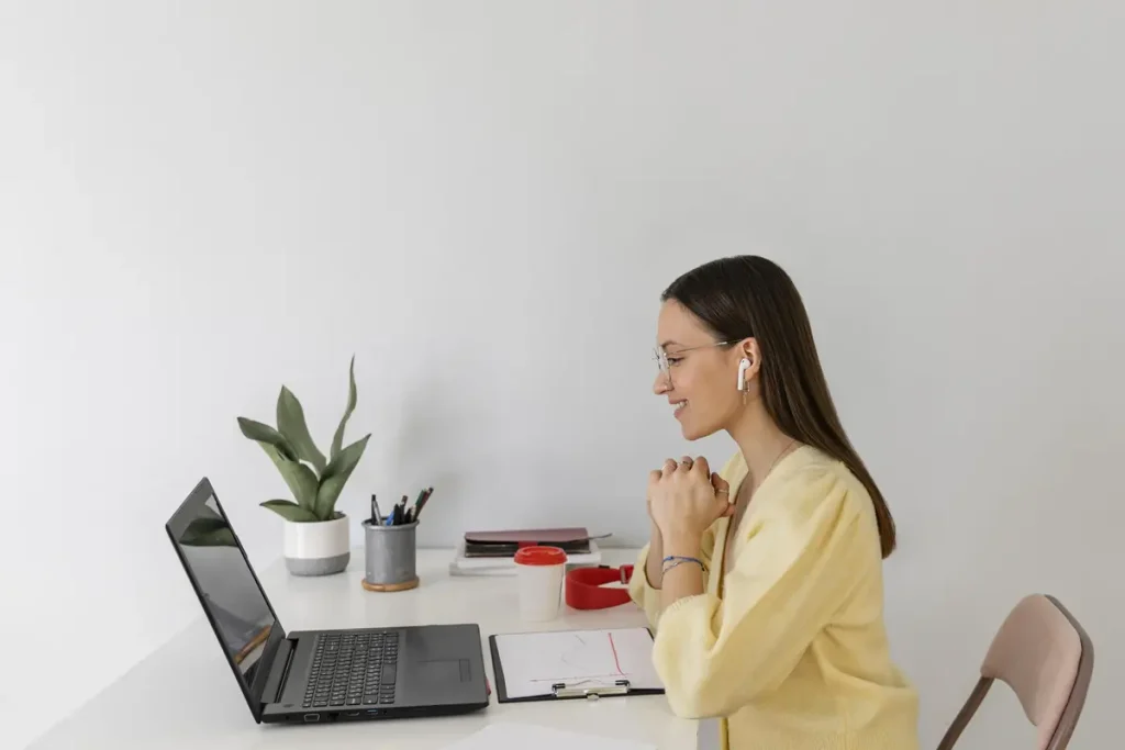 Professional virtual assistant wearing earbuds and smiling while working on a laptop at her desk.